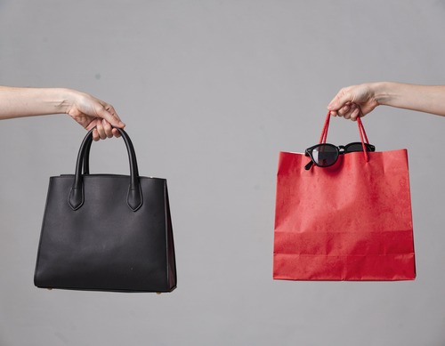 Hands Holding Black Handbag and Red Shopping Bag with Sunglasses on Gray Background